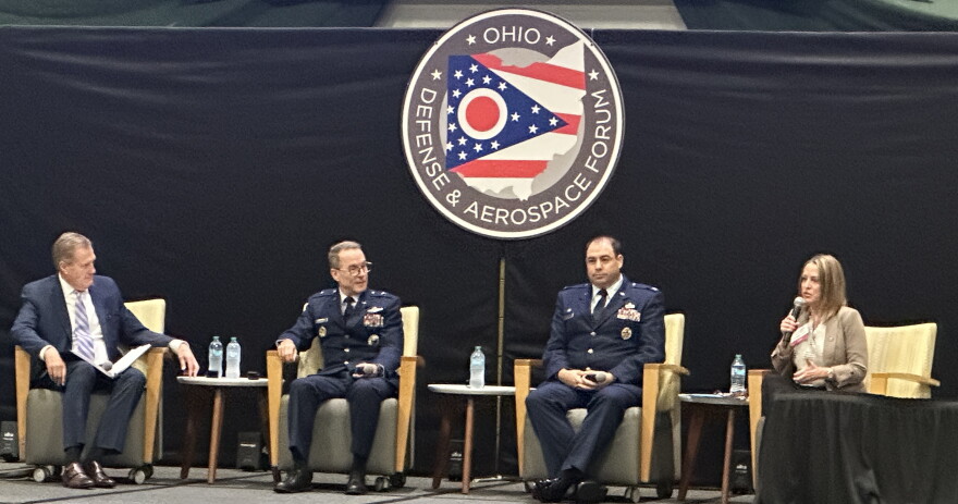 Three men and a woman sit in chairs on a stage below an an Ohio Defense & Aerospace Forum sign.