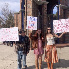 Three high school students hold signs in front of the Santa Cruz Clocktower. From left to right they read: "We are skipping our lesson to teach you one," "Preach love not hate!" and "When cruelty becomes normal compassion becomes radical".