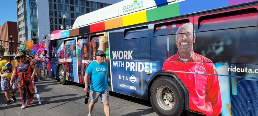 A UTA bus with the colors of the progress pride flag across the top. It also has pictures of various people smiling and the phrases "work with pride" and "move with pride."