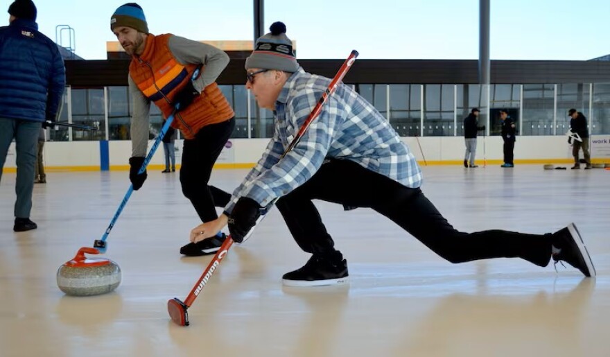 Two men play curling on an outdoor ice rink, with one sliding forward in a low lunge while releasing a granite stone and the other sweeping the ice ahead of it with a broom.