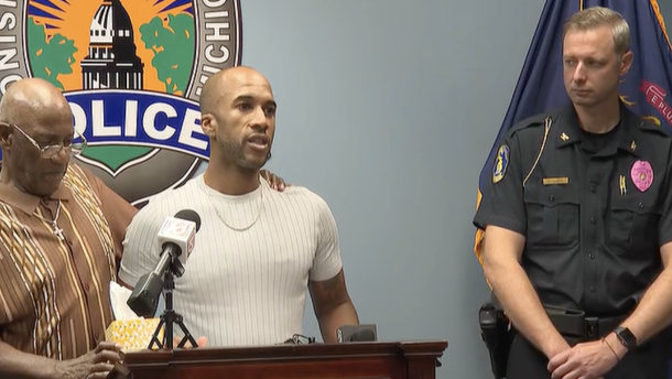 Two men stand at a podium and speak into a microphone next to a police officer