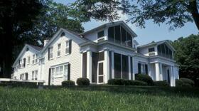 A large, two-story white historic house sits on a grassy hill under a clear blue sky. The building features multiple gabled rooflines, dark shutters on its windows, and several screened-in porches with large white columns. Lush green trees frame the sides and top of the image, casting soft shadows on the lawn in the foreground