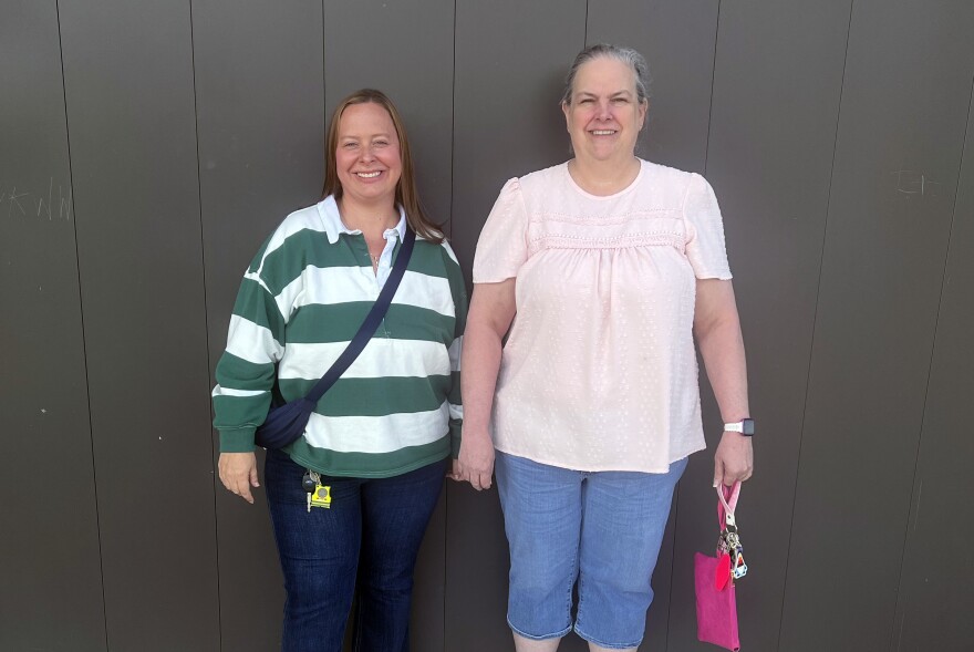 Two women stand next to each other in front of a brown wall.