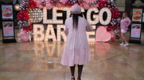  A woman wearing a pink dress takes a picture of a sign that says "Let's Go Barbie."