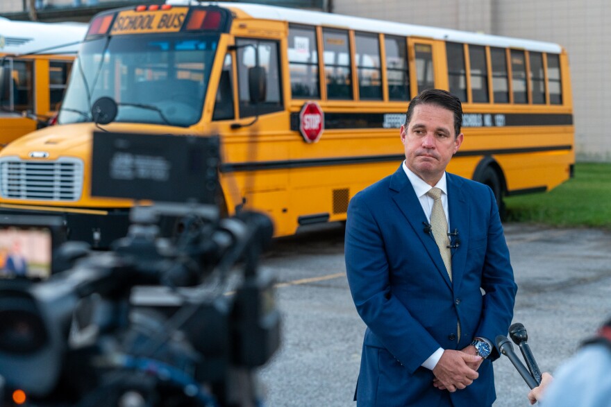 A man in a suit stands before a TV camera and a collection of microphones. He is in a parking lot. A school bus is parked in the background.