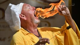 A man wearing a yellow peasant shirt and a white chef's hat places a flaming torch inside his mouth.