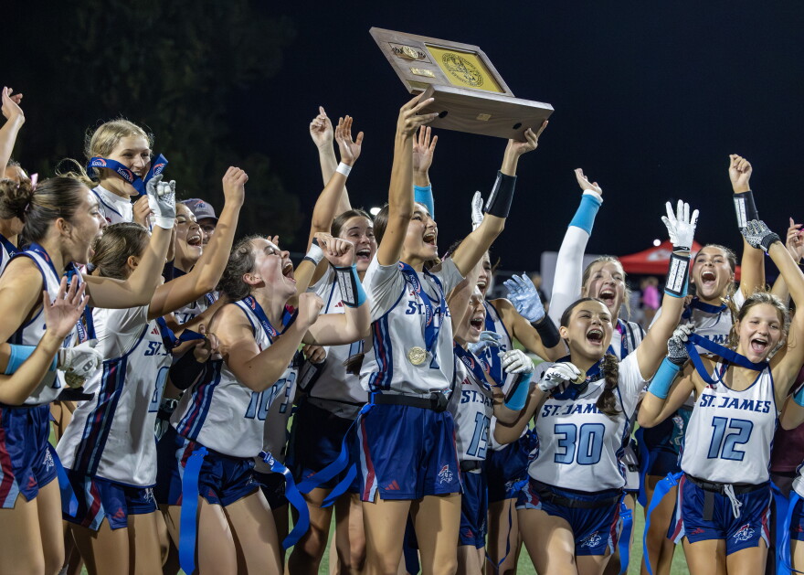 Quarterback Emme Albright is lifting up the plaque declaring them state champions alongside her St. James team. They are all in their blue and white uniforms and many of them have medals around their necks and in their hands showing off the win. They are jumping in celebration.