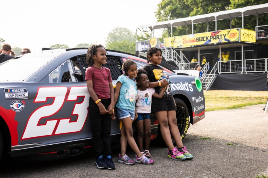 Four small Black children stand smiling in front of Bubba Wallace's race car, adorned with McDonalds advertising and the number 23