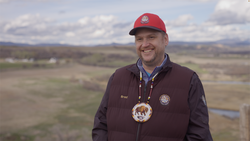 A man with a short brown beard wears a red hat  and brown jacket. He smiles and stands in front of a field. 