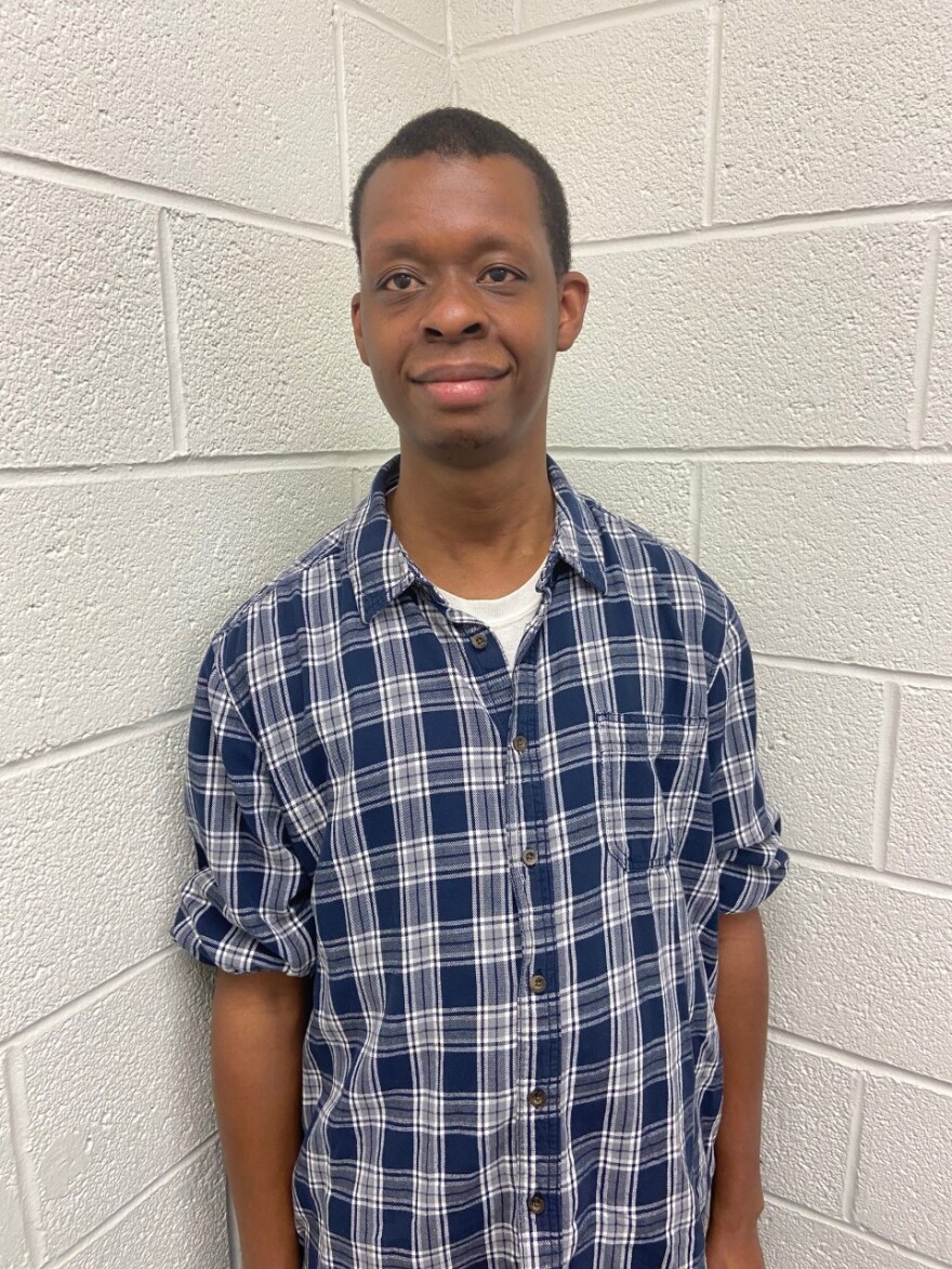 Terrance Hasan smiles at the camera, wearing a blue plaid button-down. He is standing in front of a white brick background.