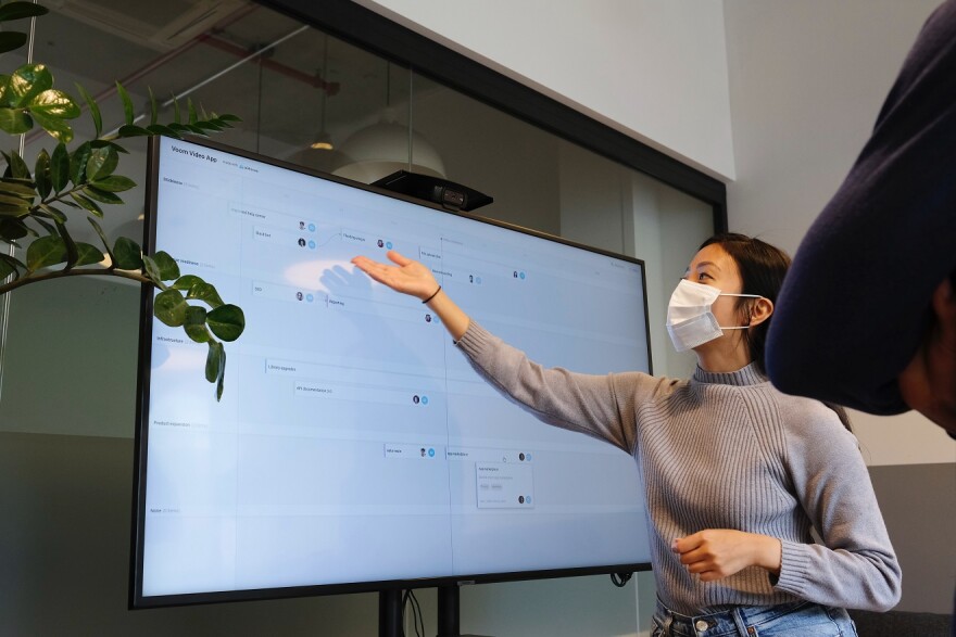 A teacher wears a mask and points to a large monitor with information on it.