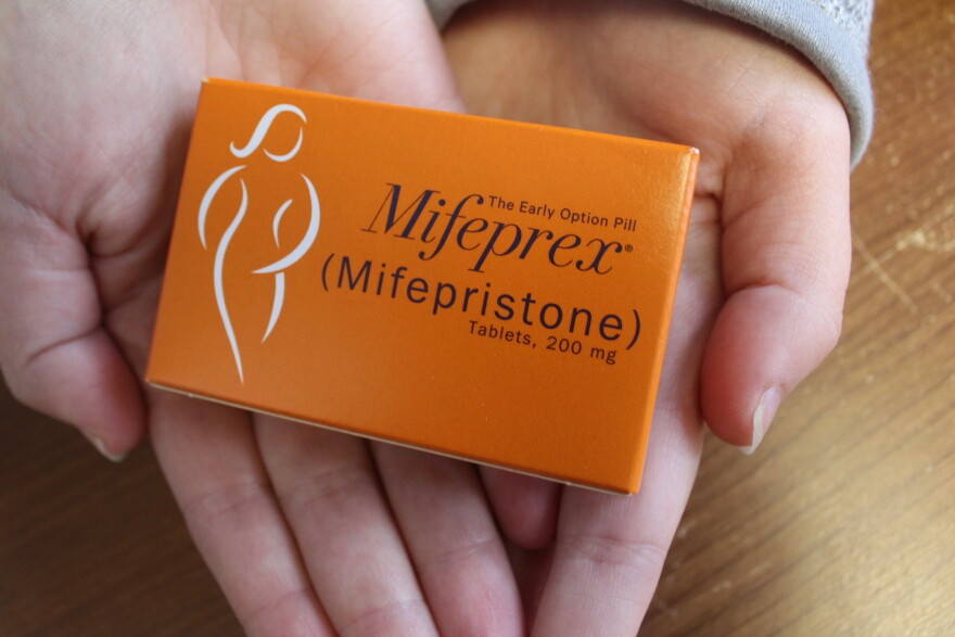 Two hands hold an orange box of pills labeled Mifeprex (Mifepristone) above a wood tabletop 