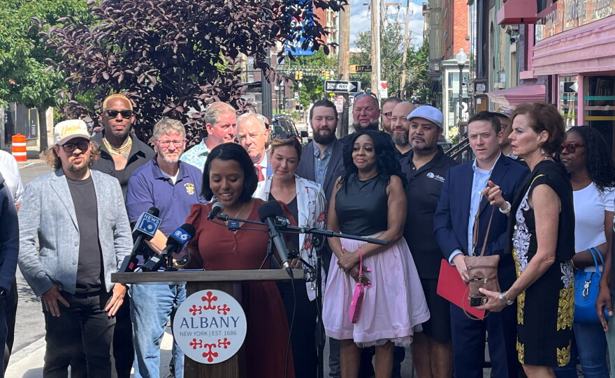 Albany Mayor Dorcey Applyrs is standing at a lectern that was placed in the sidewalk. This meant that people had to cross the street to get around the press conference. There's a bunch of people standing behind her. 