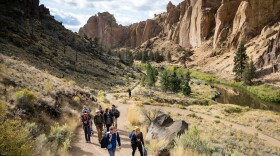 About eight people climb a wide sandy trail, with rock cliffs in the background.