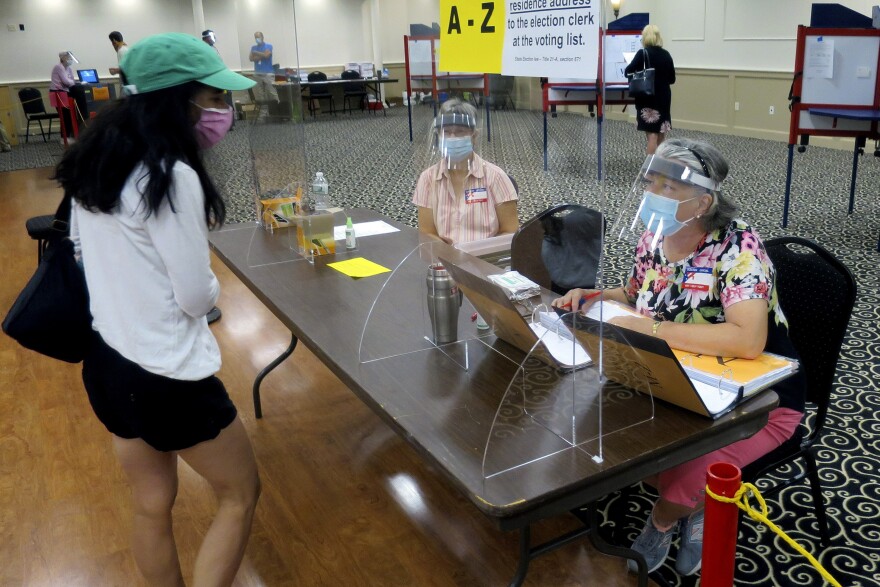 Election workers Adonlie DeRoche, seated left, and Judy Smith, seated right, wear masks and face shields and work behind plexiglass for safety during the coronavirus pandemic, while assisting a voter during primary elections on Tuesday, July 14, 2020, in Portland, Maine. Voters were encouraged to vote ahead of time via absentee ballot, but polling stations were available for in-person voting. (AP Photo/David Sharp)