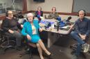 Five people wearing headphones sit at a table in a radio talk studio: a woman front left has short brown hair and is wearing glasses, a black top and grey pants; a woman front right has short grey hair and is wearing a turquoise blazer, turquoise skirt, white shirt and black flats; a woman back left has short grey hair and is wearing a black blazer, purple shirt and white clerical collar; a man back right has short brown hair and is wearing a green fleece over a green button-down shirt; a man at center has short dark hair and is wearing a blue button-down shirt, jeans and sneakers.