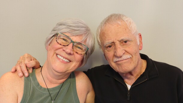 Kay and Arnold Martinez share a sideways hug at their StoryCorps appointment in St. George, Utah.  Kay has short white-gray hair and wears glasses with colorful frames, blue and black on the top tan and black on the bottom.  She smiles with one arm under her father's arm. She is wearing an army green tank top, and a large green pendant hanging from a dark cord around her neck.  Arnold has one hand on Kay's shoulder, and his thumb is holding his index finger.  Arnold has white hair and mustache and wears a black  pullover with a short zipped placket, a left chest pocket with a button, an da piece of paper  slightly extending from the pocket.  Arnold looks toward the camera in a matter-of fact way, his dark eyes expressing love, and his mouth in a straight line.