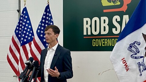 a man speaks into microphones next to American and Iowa flags