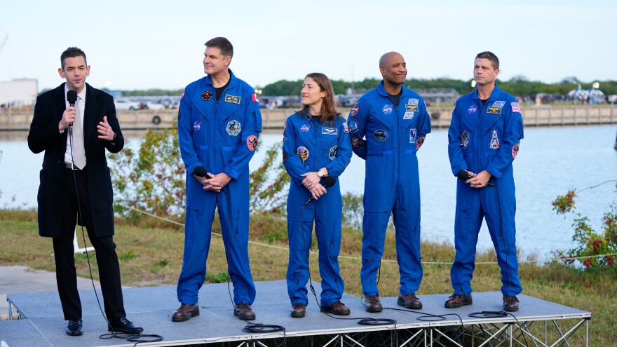 A man in a suit and holding a microphone speaks, while four people to the right wearing blue jumpsuits look at him. They're outdoors with a lake behind them.