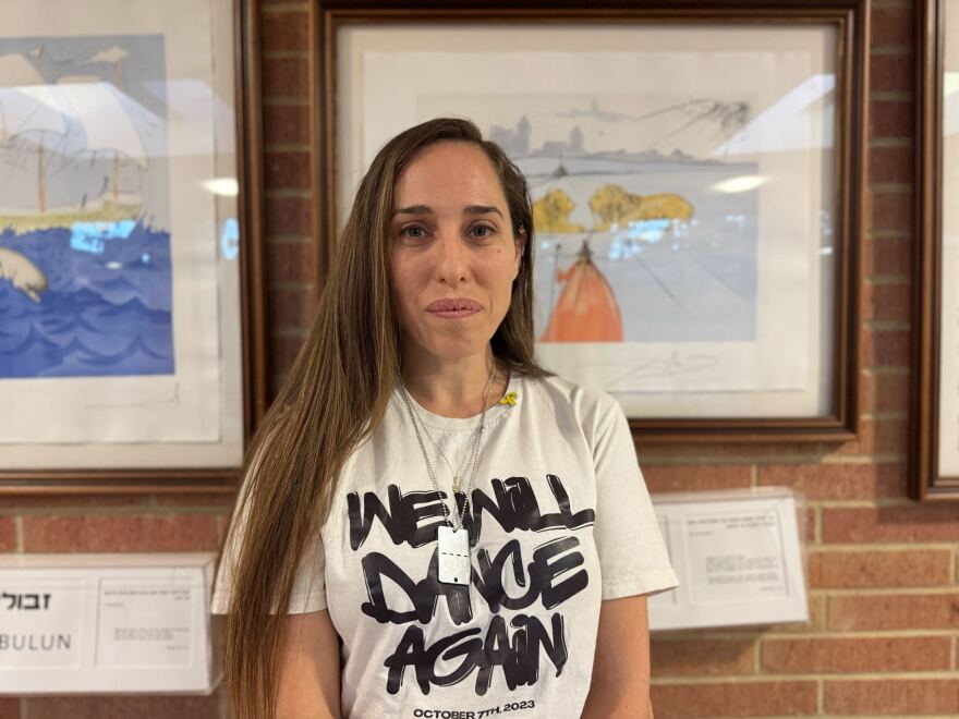 A woman in a white shirt with "We will dance again" in black lettering stands against a brick wall with framed art on it.