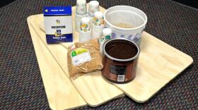 Three pine boards are lying on the carpet. The boards are 1/2 "plywood, approximately 1.5 feet by 2 feet. There are five stacks of bathroom cups, a gallon bucket of sand, a box of table salt, a gallon can of coffee, and a bag of flaxseed on top of the boards.