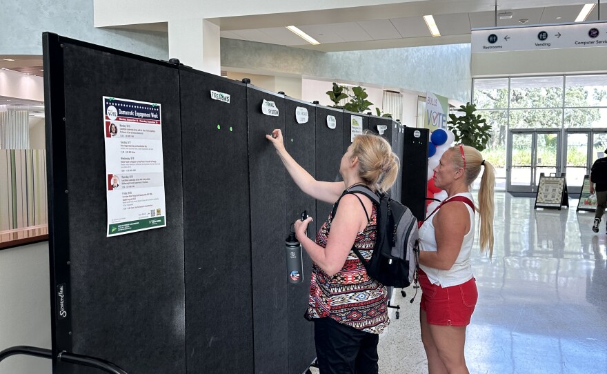 Two women are standing in front of a tall black felt board. The board is separated into columns representing issues that voters may care about, such as 'reproductive rights' and 'the economy'. One of the woman is putting a sticker on one of the columns.