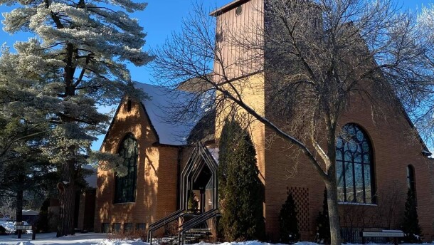 A stately brick church building in the winter with frost on surrounding pine trees against a bright blue sky.