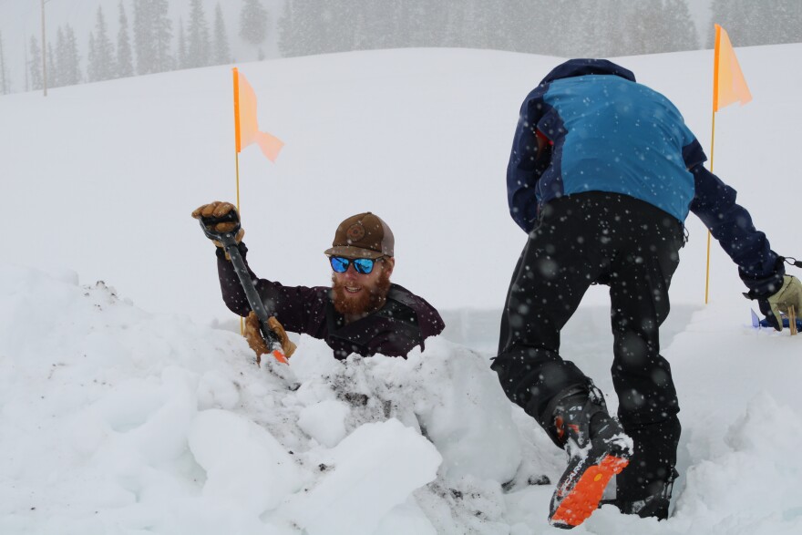 A man stands neck-deep in a snow pit, pulling out a shovel while snow falls