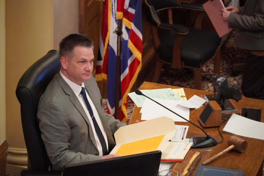 Man in suit and tie sits before desk and gavel, holding official papers and listening as someone else out of frame speaks.
