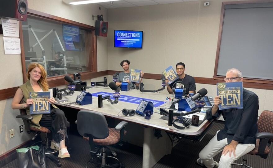 Four smiling people holding copies of CITY Magazine as they sit around a table in a radio talk studio: a woman front left has long wavy red hair and is wearing a green top, black pants, and white sandals; a man back left has short curly brown hair and is wearing a gold chair and a blue graphic t-shirt; a man back center has short dark hair and is wearing glasses, a gold chain, and a black graphic t-shirt; a man at right has very short dark hair and is wearing a black button-down shirt, tan linen pants, and grey and blue sneakers