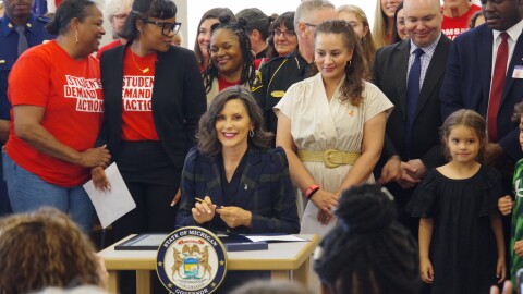 Governor Whitmer looks at the camera and smiles as she sits at a table with the seal of Michigan on the front. She is holding a pen. To the right of her are two children, and she is surrounded by people. 