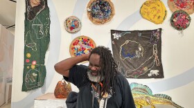 Artist Paul Chandler smiles while standing in front of a display of his mixed-media artwork at the Buffalo AKG Art Museum. The wall behind him is filled with colorful embroidered and beaded textile pieces, alongside sculptures on pedestals. 