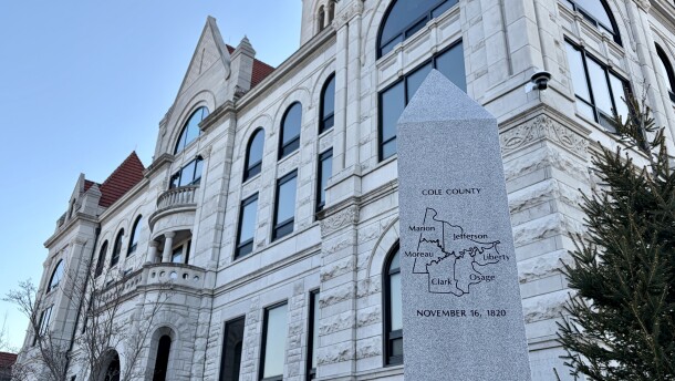 The white brick, gothic architecture Cole County courthouse exterior is in the background. A pillar with a map of Cole County is in the foreground. 