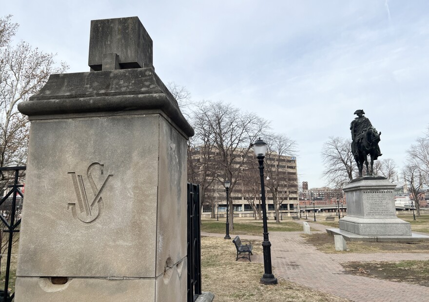 Outdoors photo on a cloudy day shows a park in wintertime. Trees are bare and there's a bronze statue of George Washington riding a horse on top of a marble pedestal. In the foreground is a concrete column that shows "WS" chiseled in it.