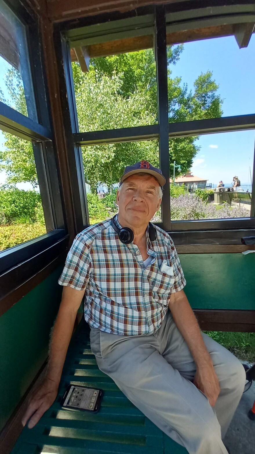 A man in light gray pants and a plaid shirt with headphones around his neck and wearing a blue baseball cap sits inside a bus stop.
