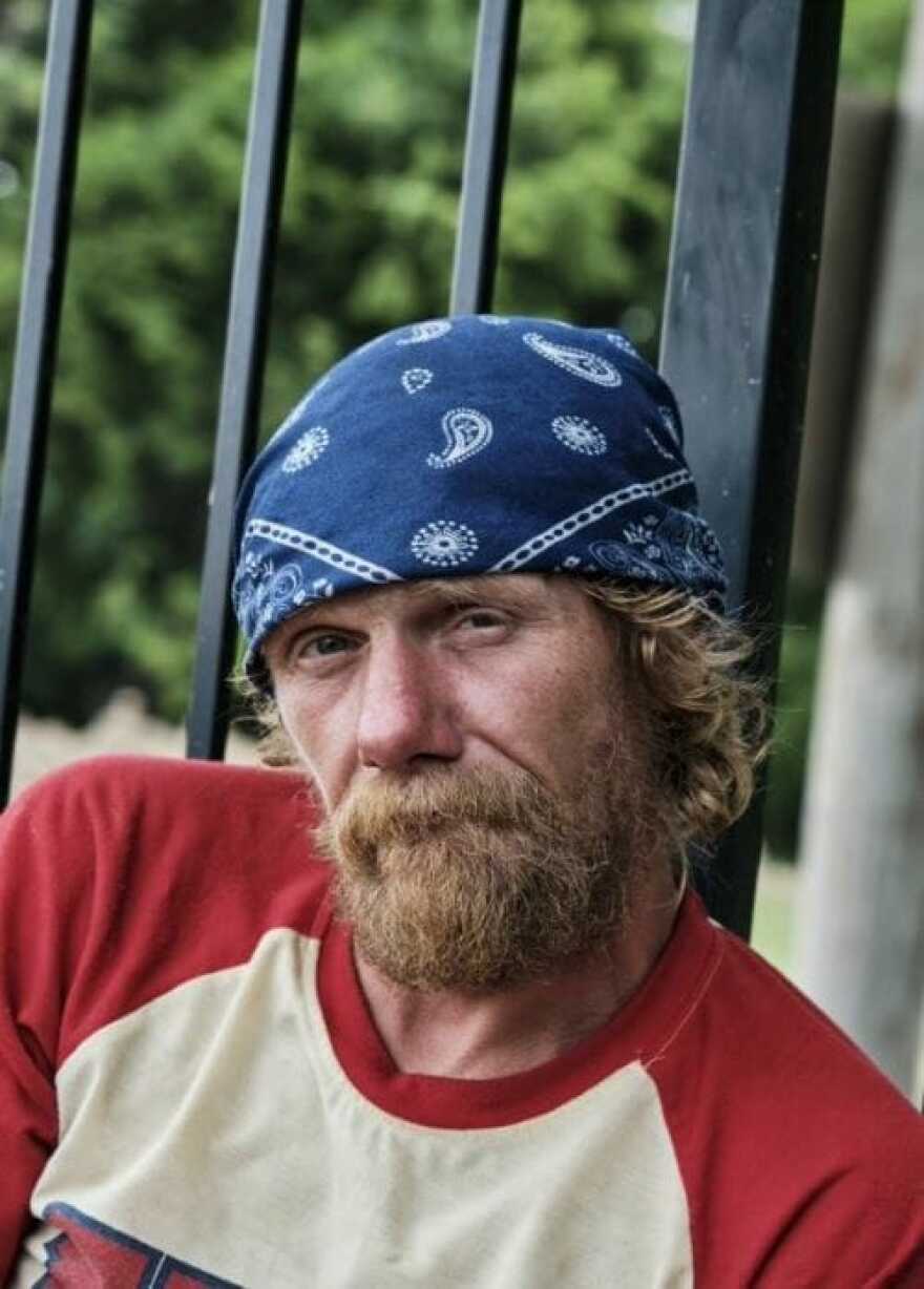 A white man with shaggy hair and a beard wears a blue bandana and a red- and cream-colored t-shirt. He looks directly ahead.