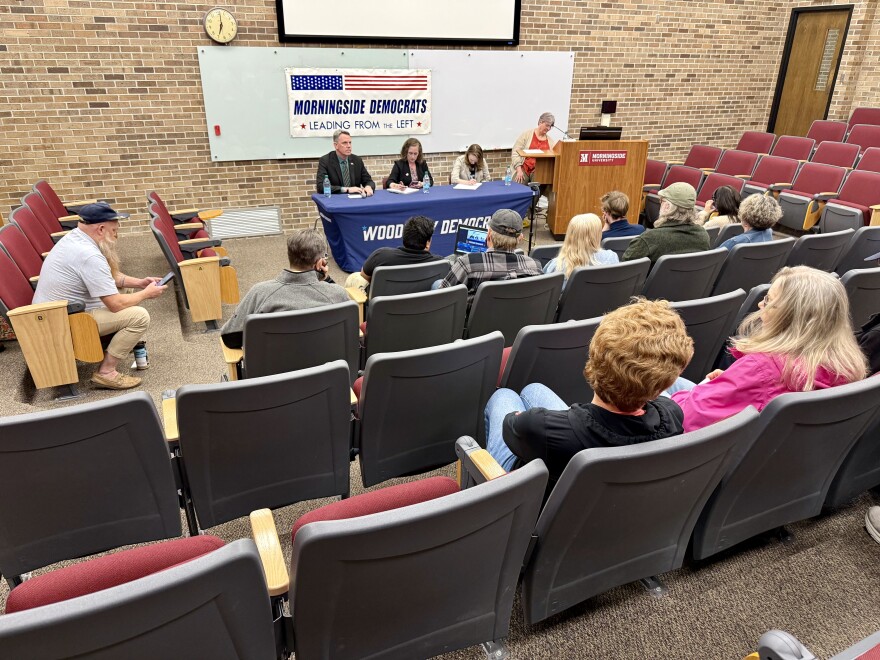 Three candidates sit in the front of a small auditorium. There is a sign behind them that says "Morningside Democrats, lead from the left."