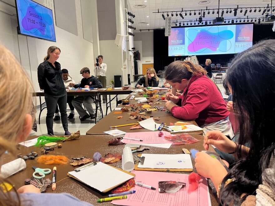 Students are working with an assortment of materials, including yarn, leaves, pencils and rocks and are focused on using the objects to make crafts. A woman is standing in front of them, looking at their work.