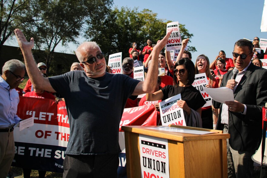 A man in a black tshirt throws his hands in the air and shrugs next to a crowd of people holding signs in favor of a union.