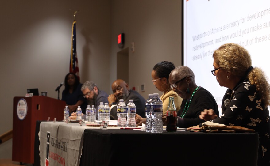 Candidates sit at a table with a projected question displayed on a screen behind them.
