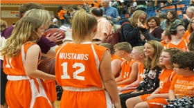 Female basketball players wearing orange jerseys standing by the bleachers