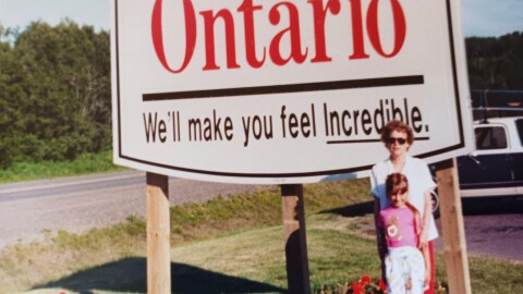 A girl and her grandma stand by a "Welcome to Ontario" sign by the side of the road.