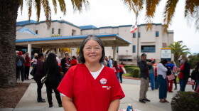 A nurse wearing red scrubs stands in front of Watsonville Community Hospital. Nurses behind her talk to one another while holding protest signs.