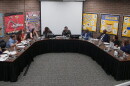 A group of people sit around a large U-shaped table in a meeting room, with colorful bulletin boards and a blank projector screen on the back wall.