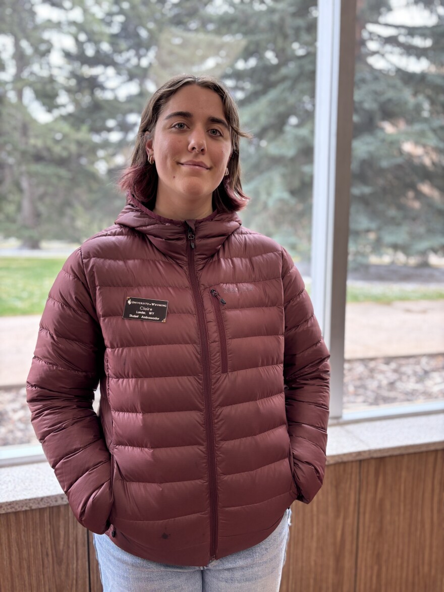 A girl wearing a purple puffy jacket with short purple-tipped hair stands in front of a window, with trees in the background.