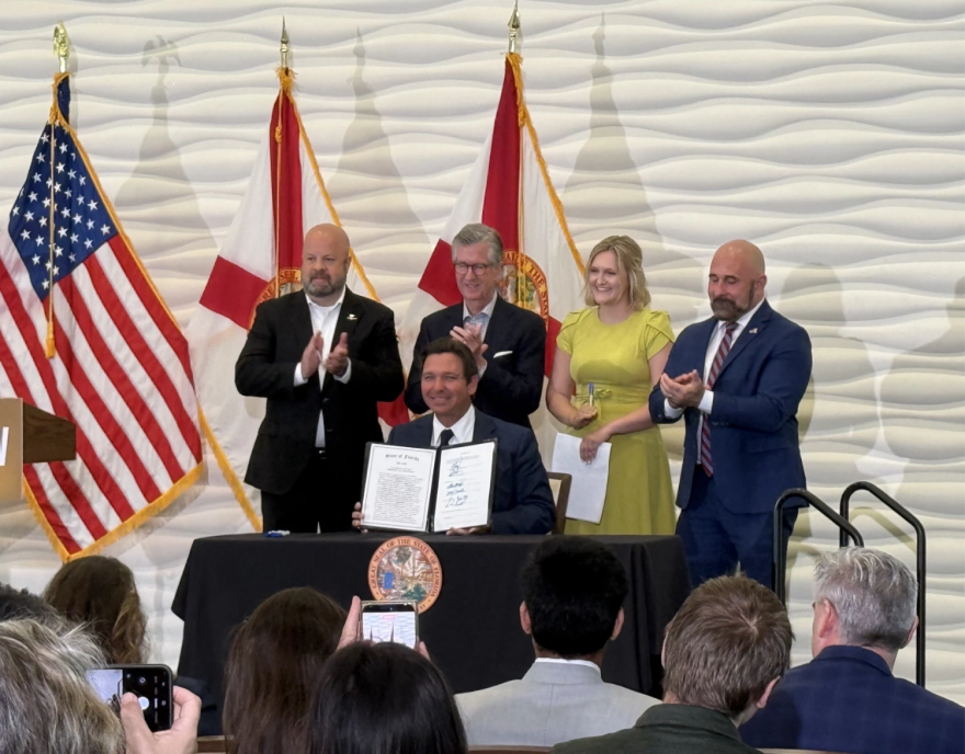 Un hombre de traje sonríe y muestra un documento a una multitud. Tres hombres de traje y una mujer con un vestido amarillo están de pie detrás de él.