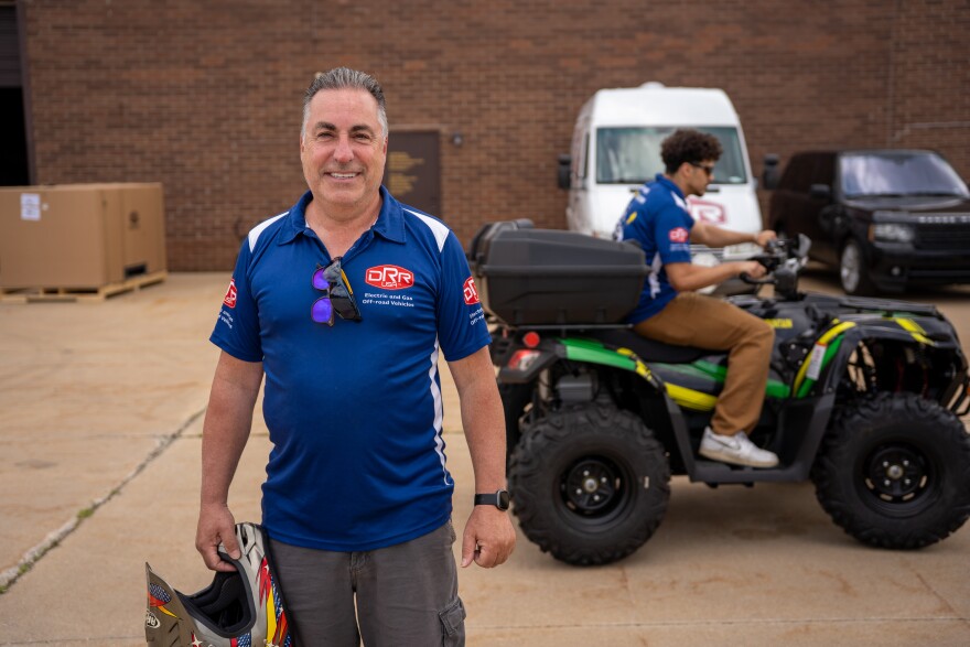Man smiles at camera while another man drives an ATV behind him