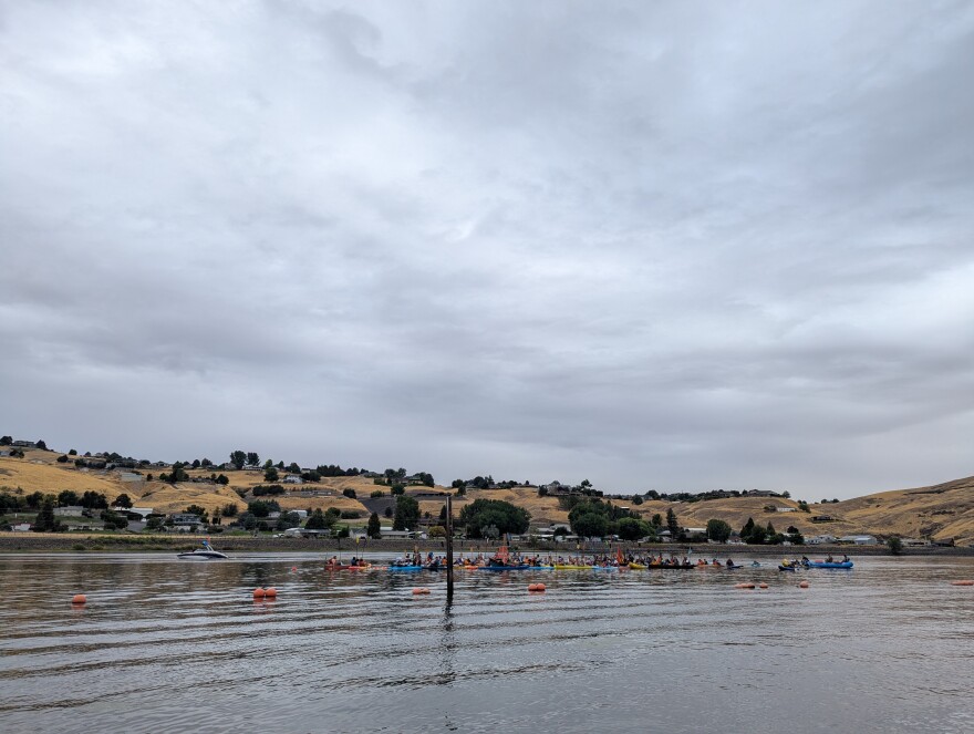 A flotilla of different watercraft float next to each other out on the waters of the Snake River. Dry hills dotted with trees can be seen in the distance under a grey sky.
