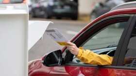 A person in a car reaches to put their ballot into a drop box.