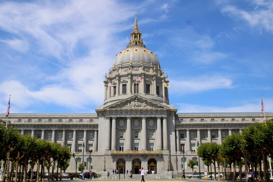 A view of the front of city hall with a few trees in the foreground and a blue sky with wispy clouds behind it. The city hall building is long and rectangular with columns in front, gold trim, and a big dome coming out of the center of it. You can see a few people walking in front of the building. 